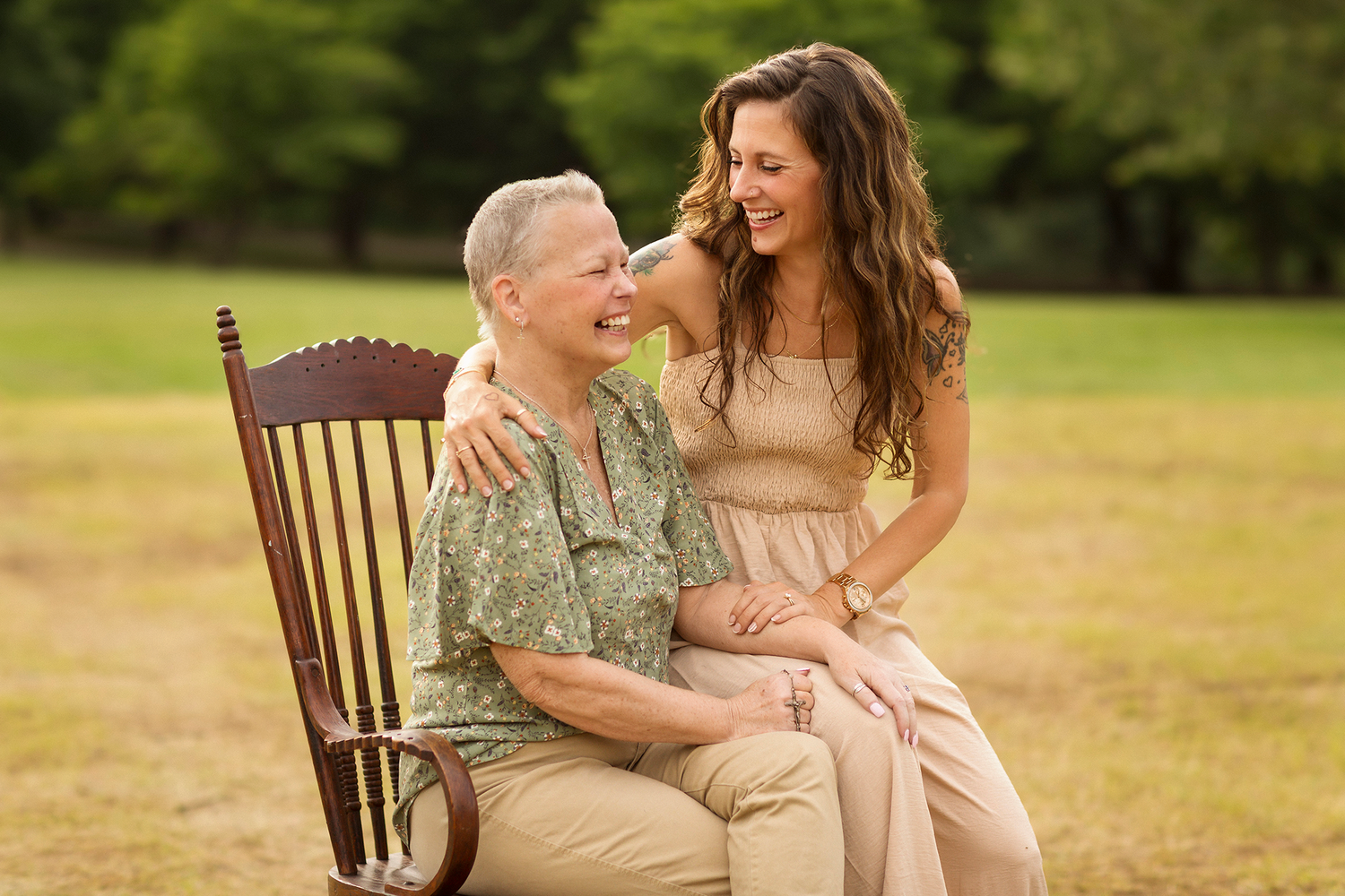 Two women sharing a tender moment while sitting together on a wooden rocking chair in a sunny outdoor field.