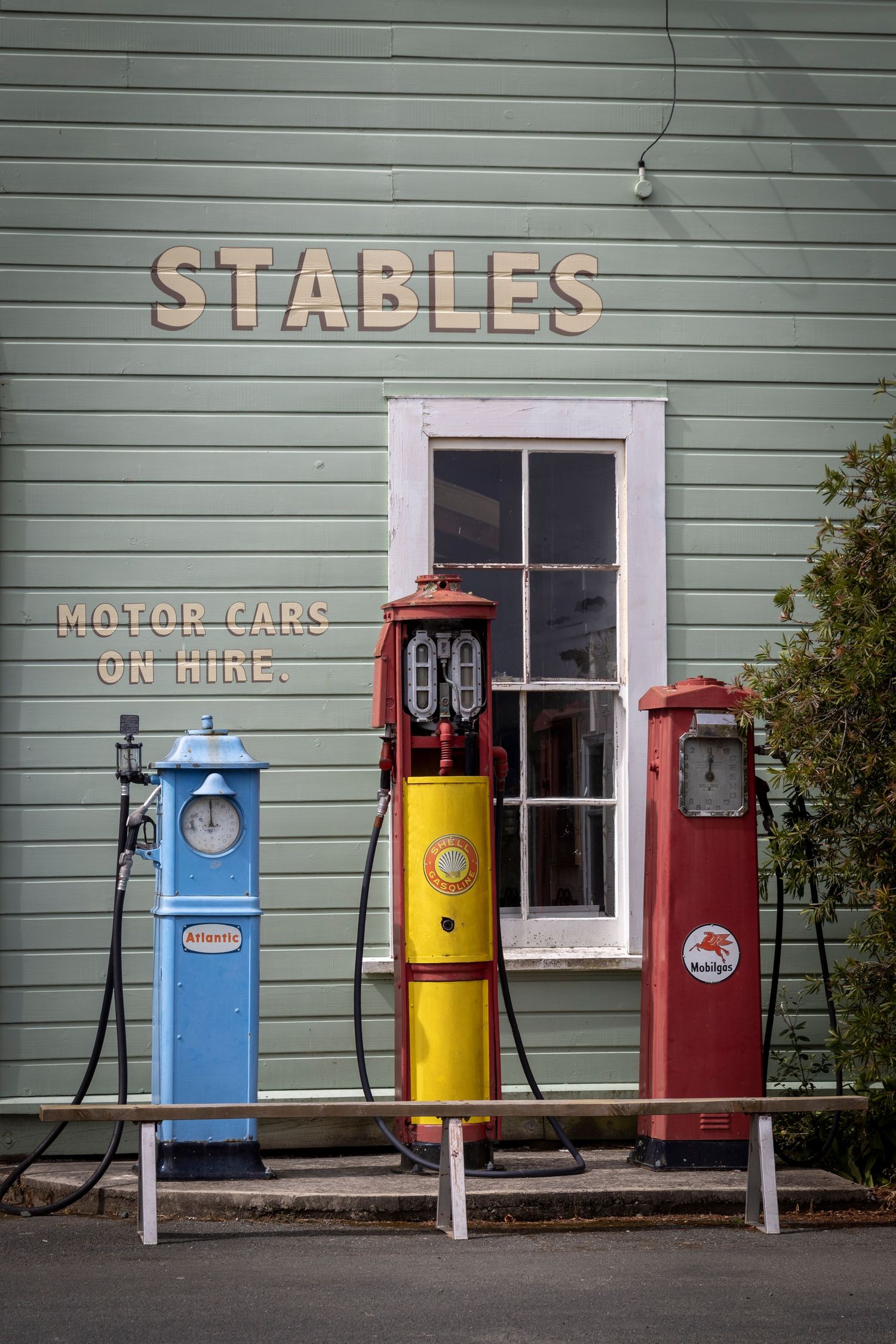 Three vintage petrol pumps—blue, yellow, and red—outside a green building labeled 'Stables' with a white-trimmed window.