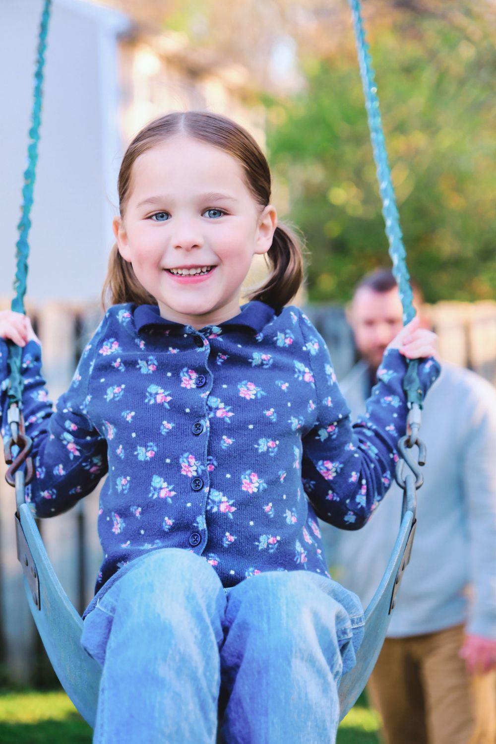 Child wearing blue floral print jacket enjoys swinging at a playground on a sunny day with blurred background.