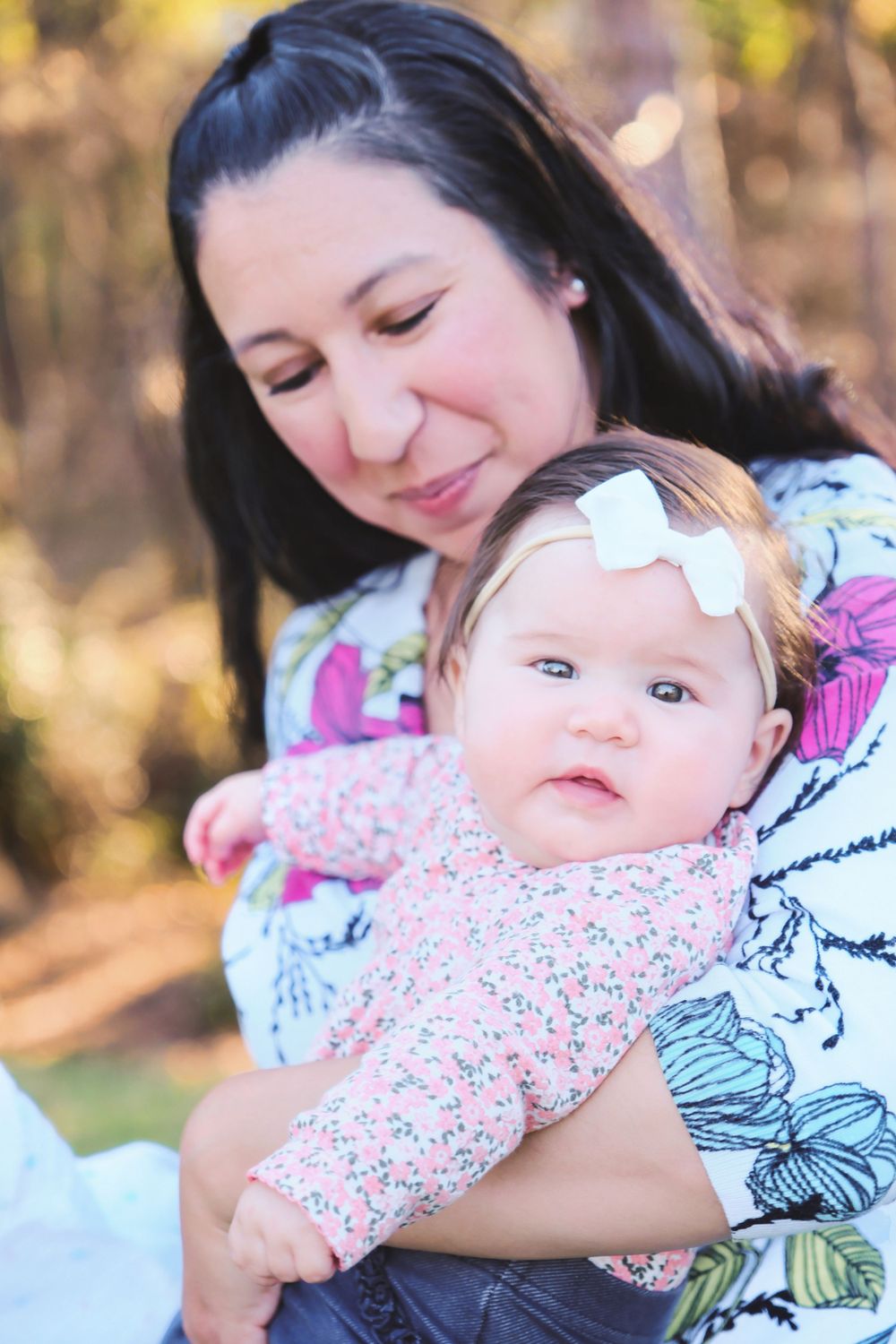 A tender moment captured outdoors as a baby wearing a floral outfit with a white bow headband is held lovingly.
