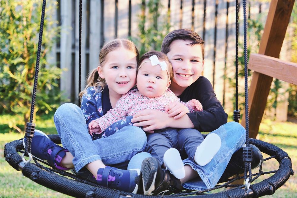 Three siblings sitting together on a round outdoor swing in a backyard garden setting.