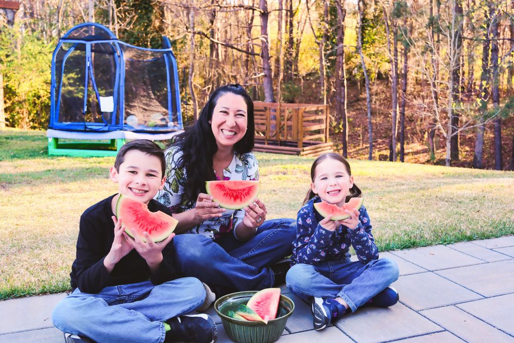 A group sits together on a patio with watermelon slices while a blue playground structure is visible in the background.
