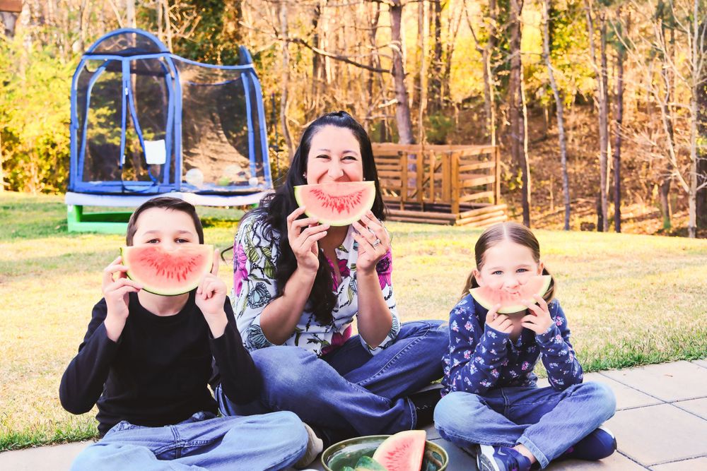 Three people sitting outdoors on concrete holding watermelon slices in front of their smiles on a sunny autumn day.