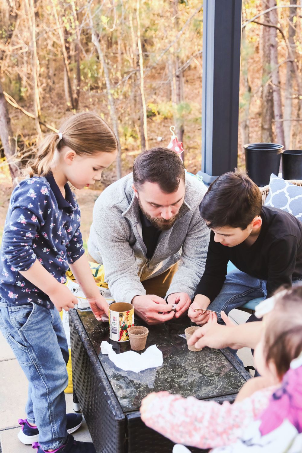 Family gathered around outdoor table engaged in crafting or activity together in wooded setting.