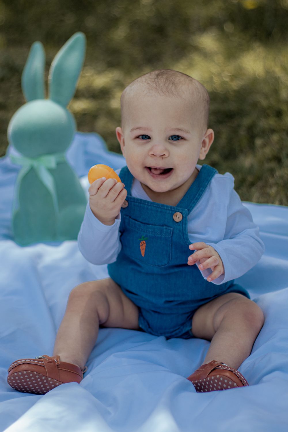 Happy baby in denim overalls sits on blue blanket with teal bunny decoration and holds an orange toy.