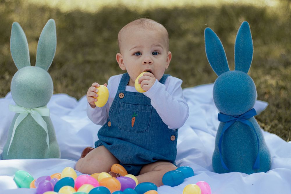Small child in blue overalls sitting on white blanket with two plush blue bunny rabbits and colorful Easter eggs.