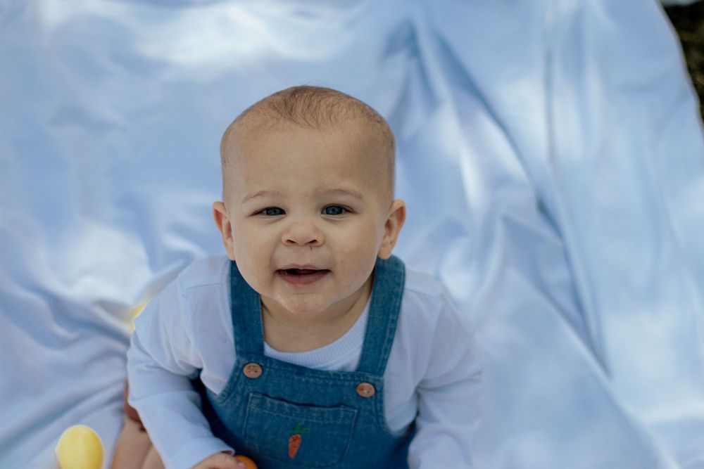 A happy baby wearing blue overalls sits against a light blue backdrop.