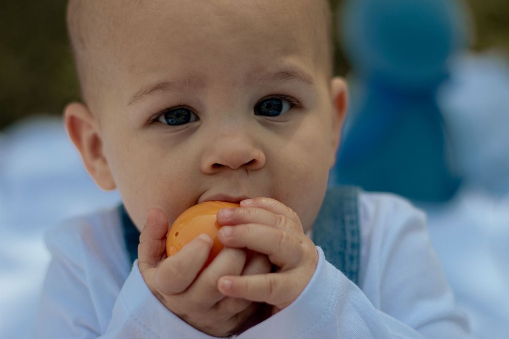 A small child takes a big bite of a fresh orange while wearing light blue clothing.