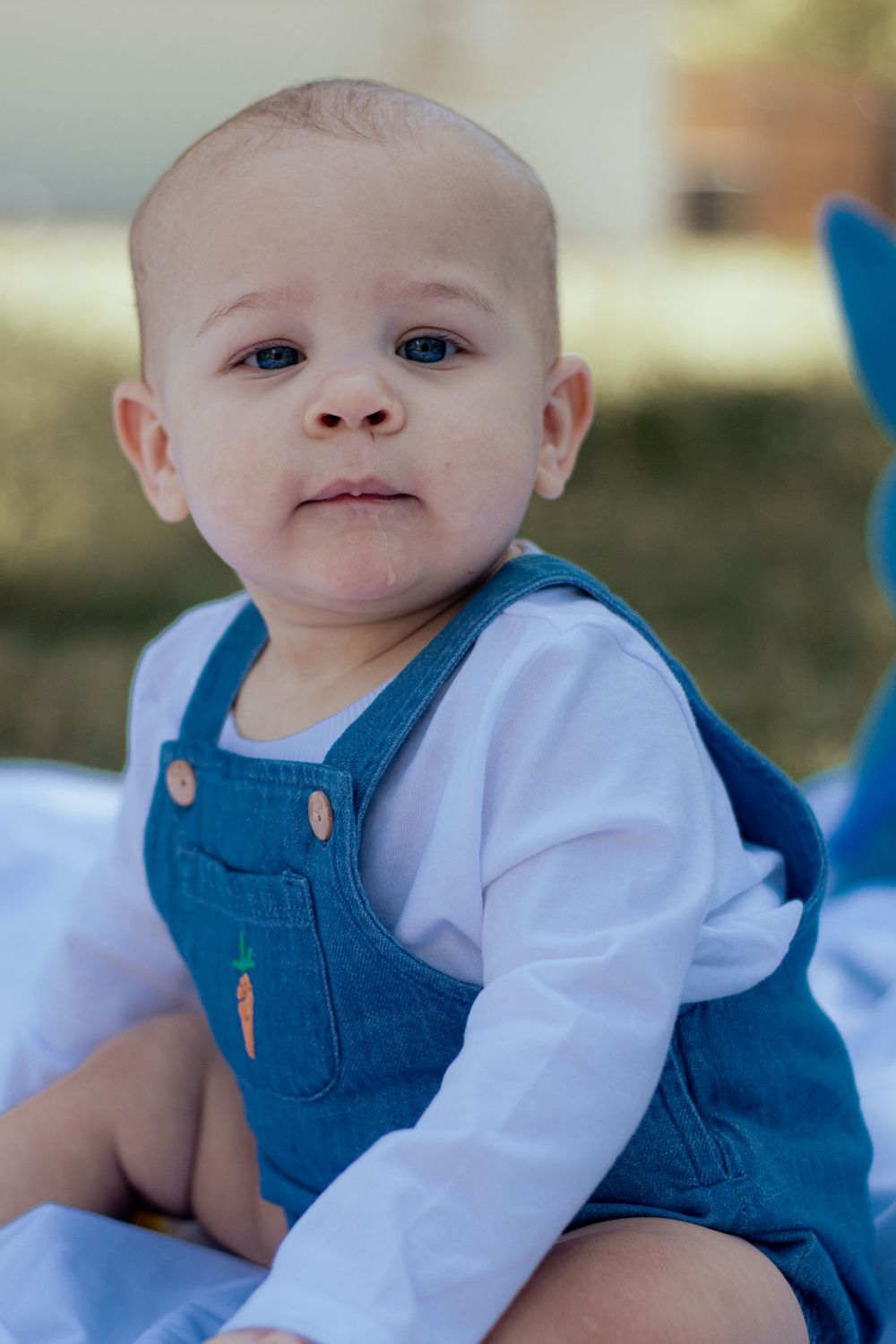 An infant wearing denim overalls and a white long-sleeve shirt sits outdoors with a gentle expression.