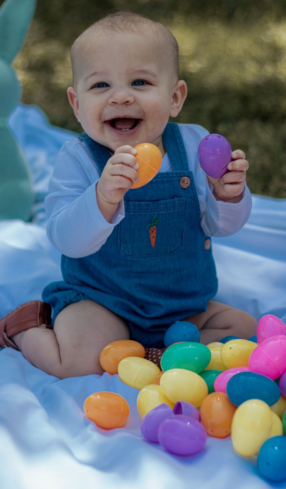 Happy baby in blue denim overalls holds colorful plastic Easter eggs while sitting on a blanket outdoors.
