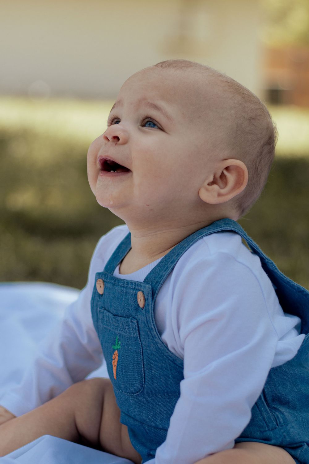 A happy baby wearing denim overalls and white shirt sits on a blanket outdoors with joyful expression.