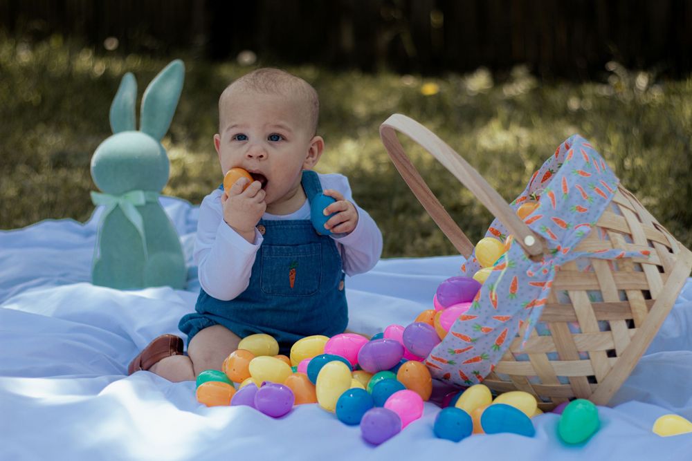 A baby wearing denim overalls sits on a blue blanket surrounded by colorful Easter eggs and a decorative bunny statue.