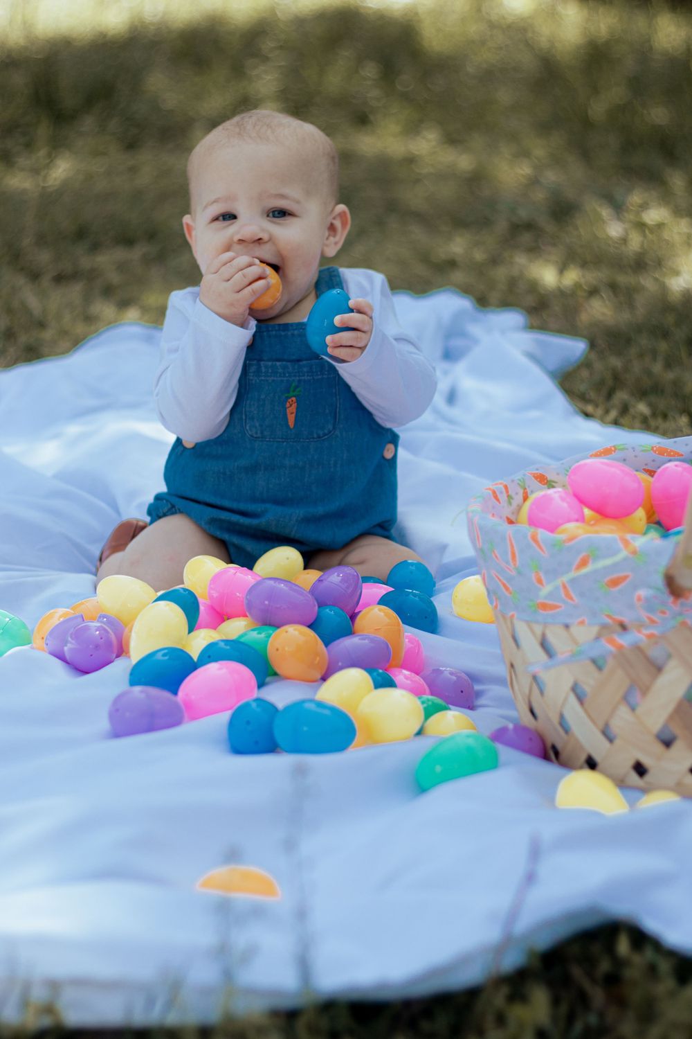Baby in blue overalls sits on blanket outdoors with colorful plastic Easter eggs scattered around and a basket nearby.