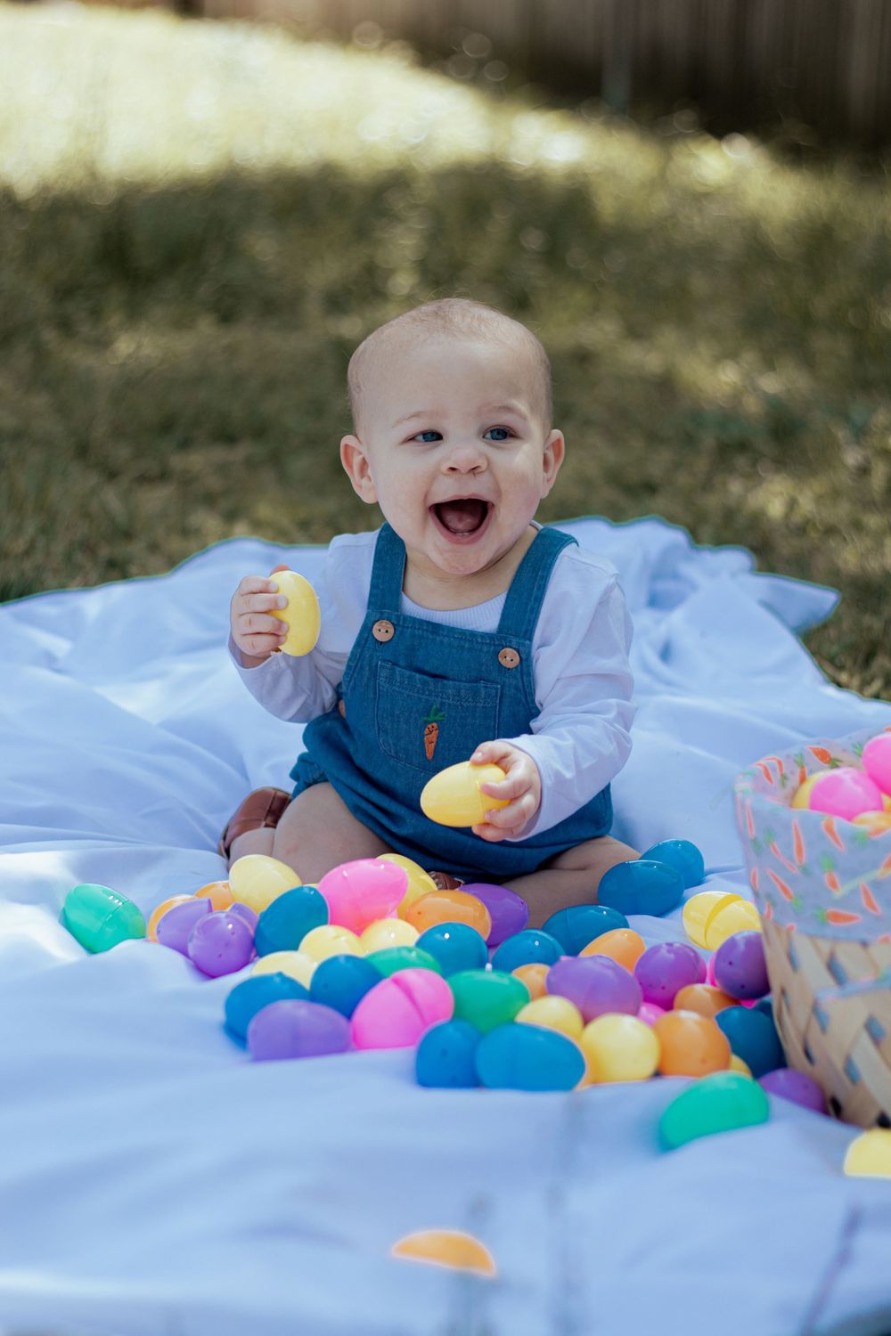 A baby wearing blue overalls sits on a blanket while playing with colorful plastic Easter eggs.