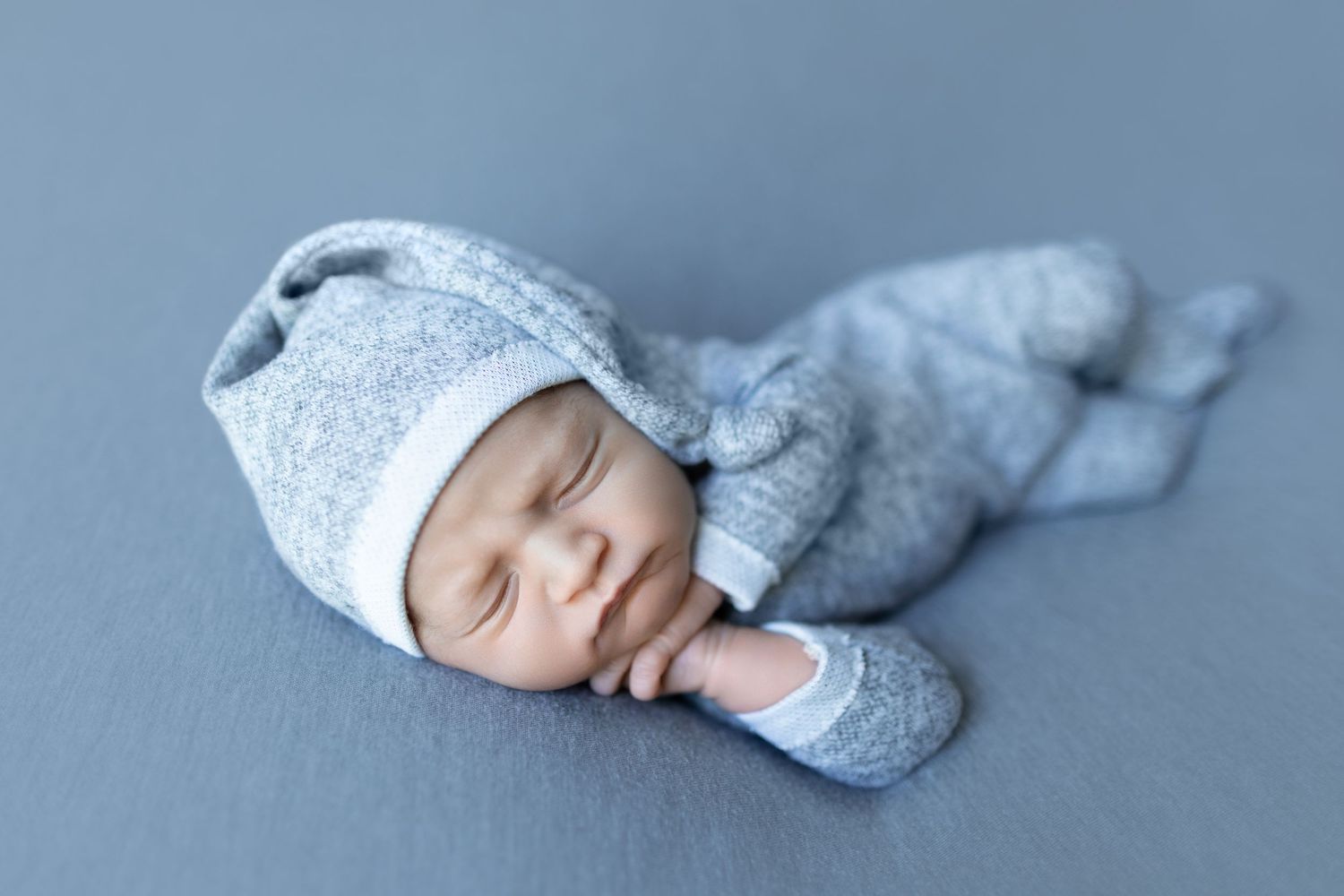 Sleeping baby wearing gray knit outfit and hat on blue backdrop.