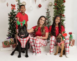 Family and two dogs wearing matching plaid pajamas pose in front of decorated Christmas trees for holiday photo.