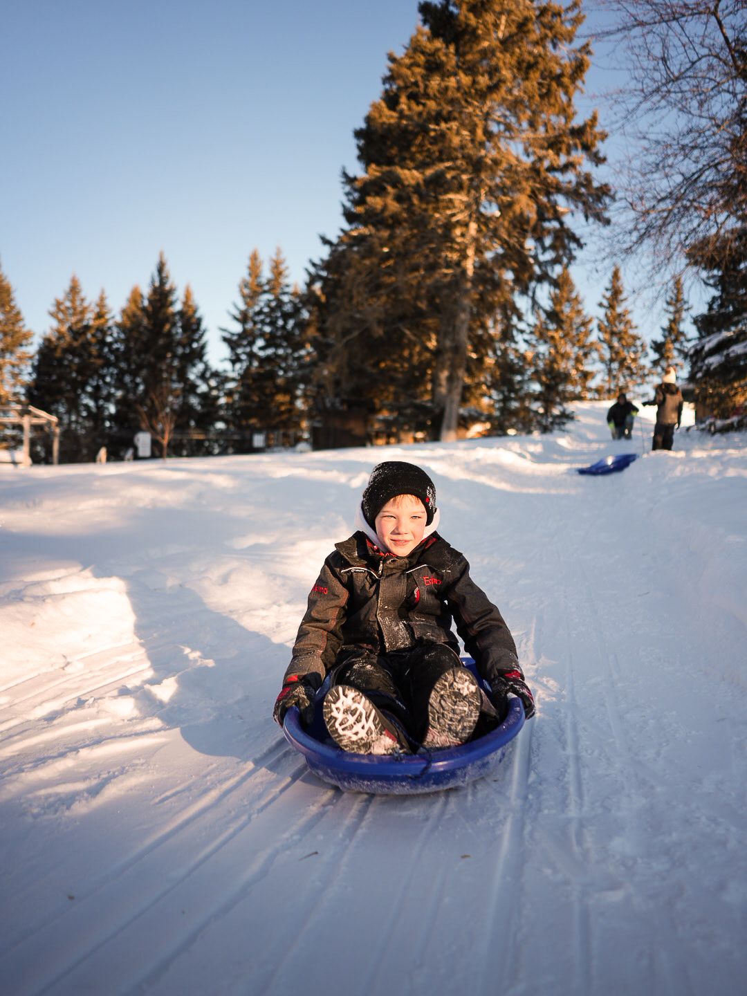 People sledding down snowy hill on blue saucer sleds at sunset.
