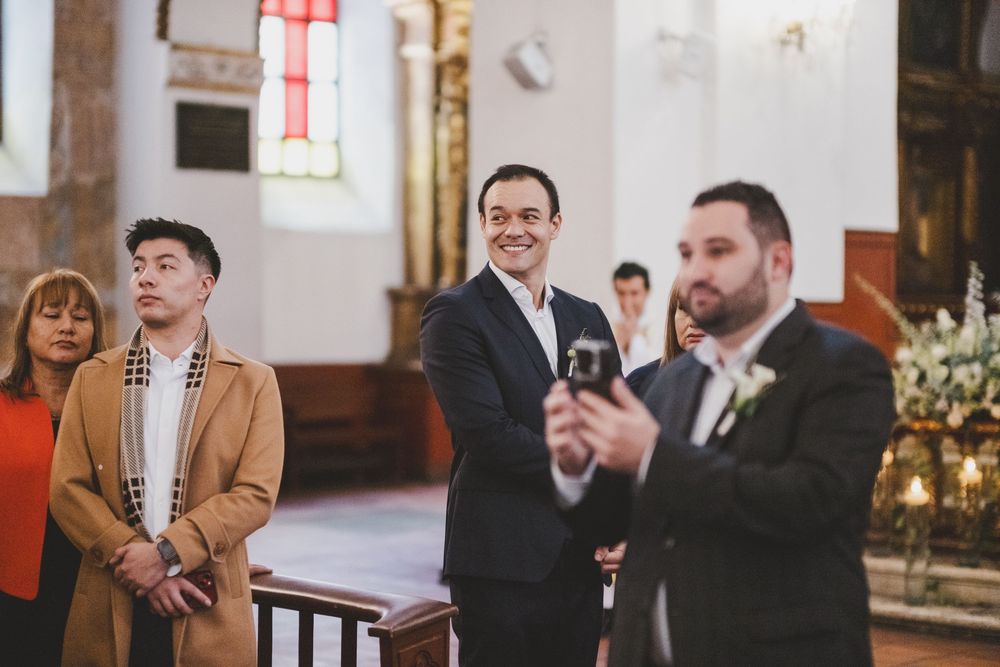 Wedding guests stand together inside a church during a ceremony with stained glass windows and floral decorations.