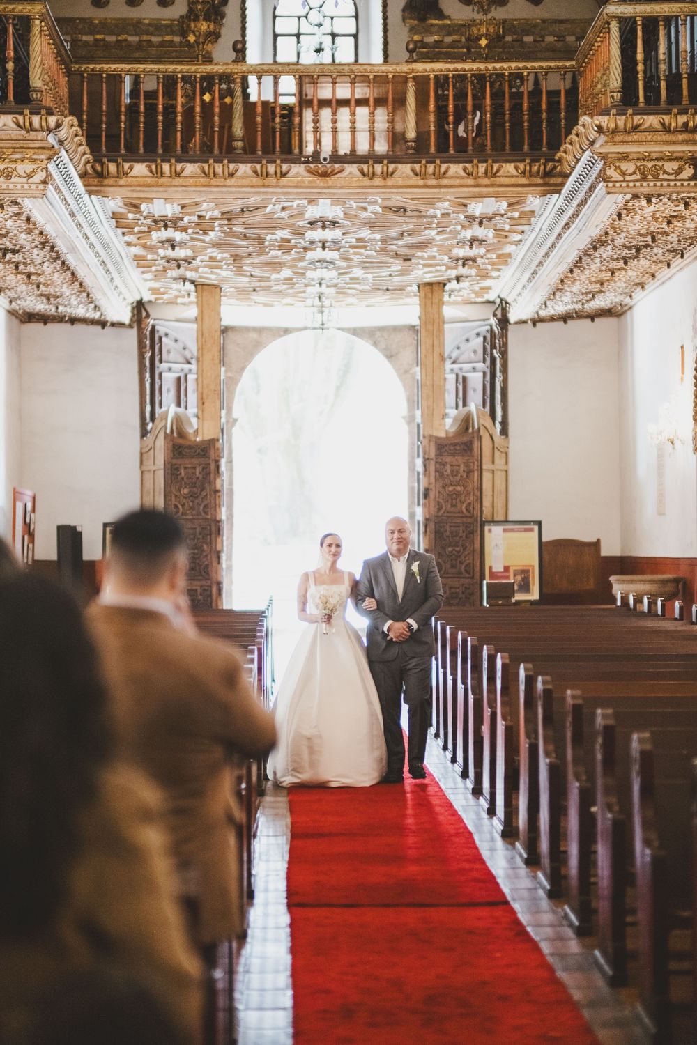 A traditional church interior with ornate wooden details and a red carpet aisle runner during a wedding ceremony.