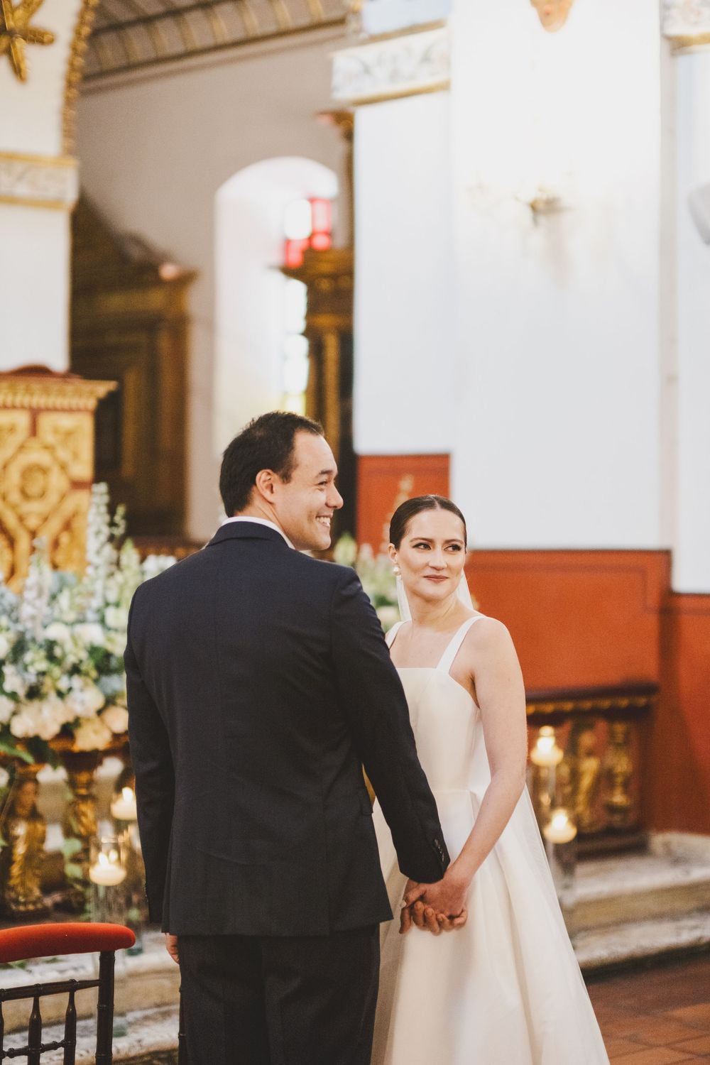 Elegant couple shares intimate moment during wedding ceremony inside ornate church with decorative columns and floral arrangements.