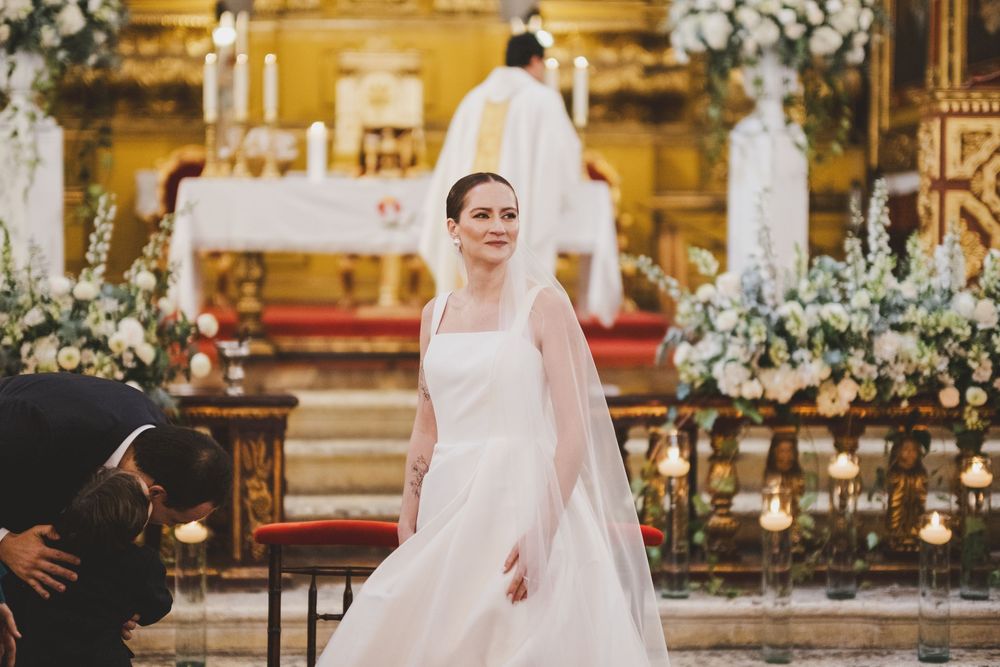 A bride in a classic white wedding gown and veil stands in an ornate church adorned with white floral arrangements.