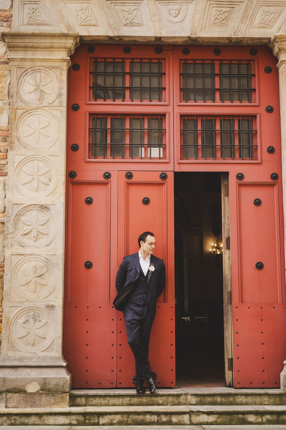 A suited figure poses in a doorway with ornate coral-colored doors and decorative stone columns.
