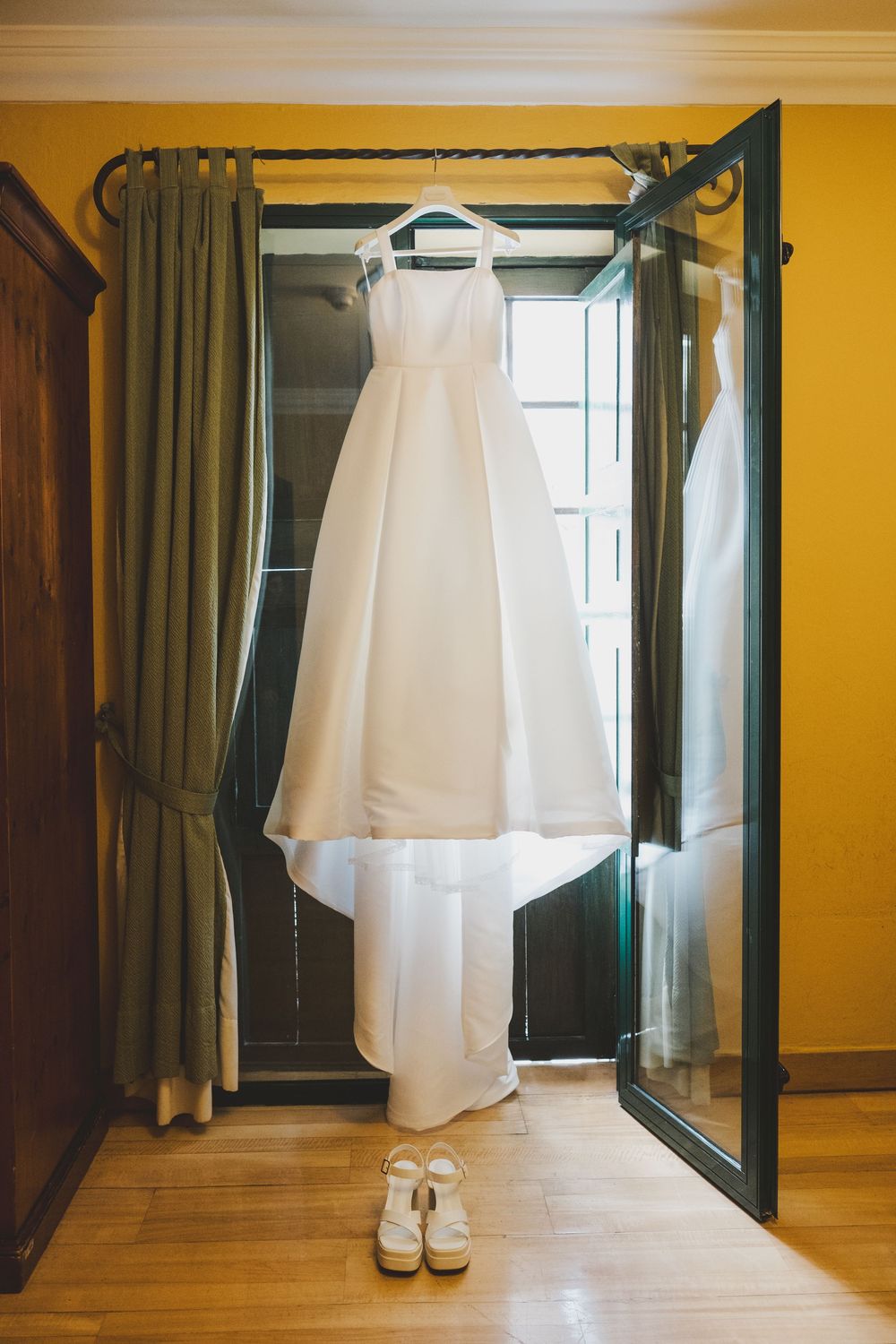 Elegant white wedding dress hangs in front of floor length mirror with white shoes beneath on hardwood floor.
