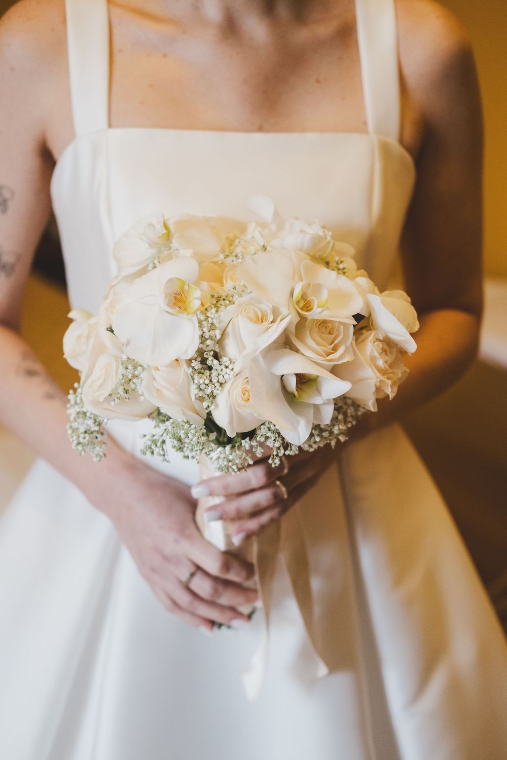 A stunning white wedding bouquet with roses and baby's breath held against an elegant white bridal gown.