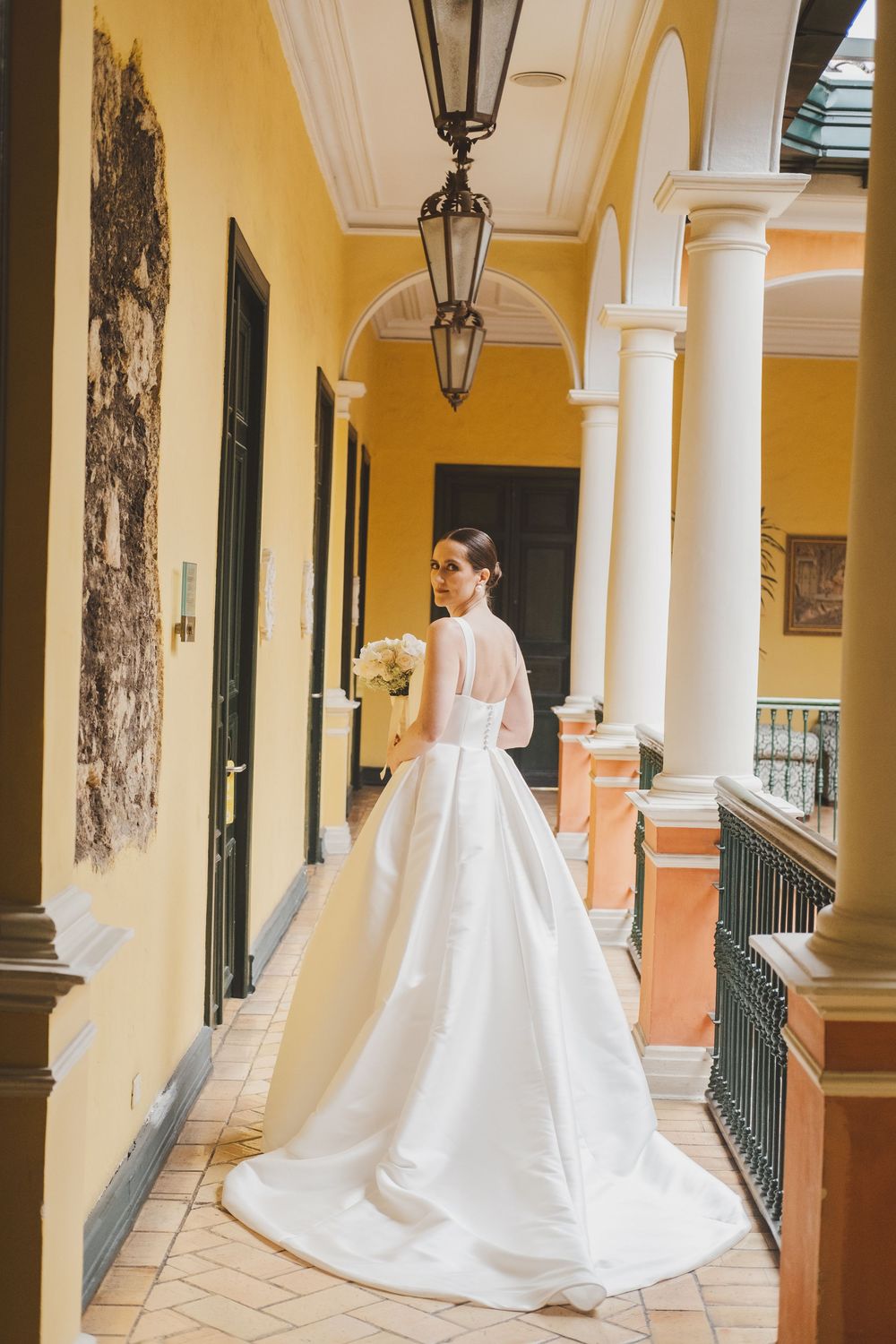 A wedding dress trailing down a yellow-walled colonnade with classic lanterns and arches.