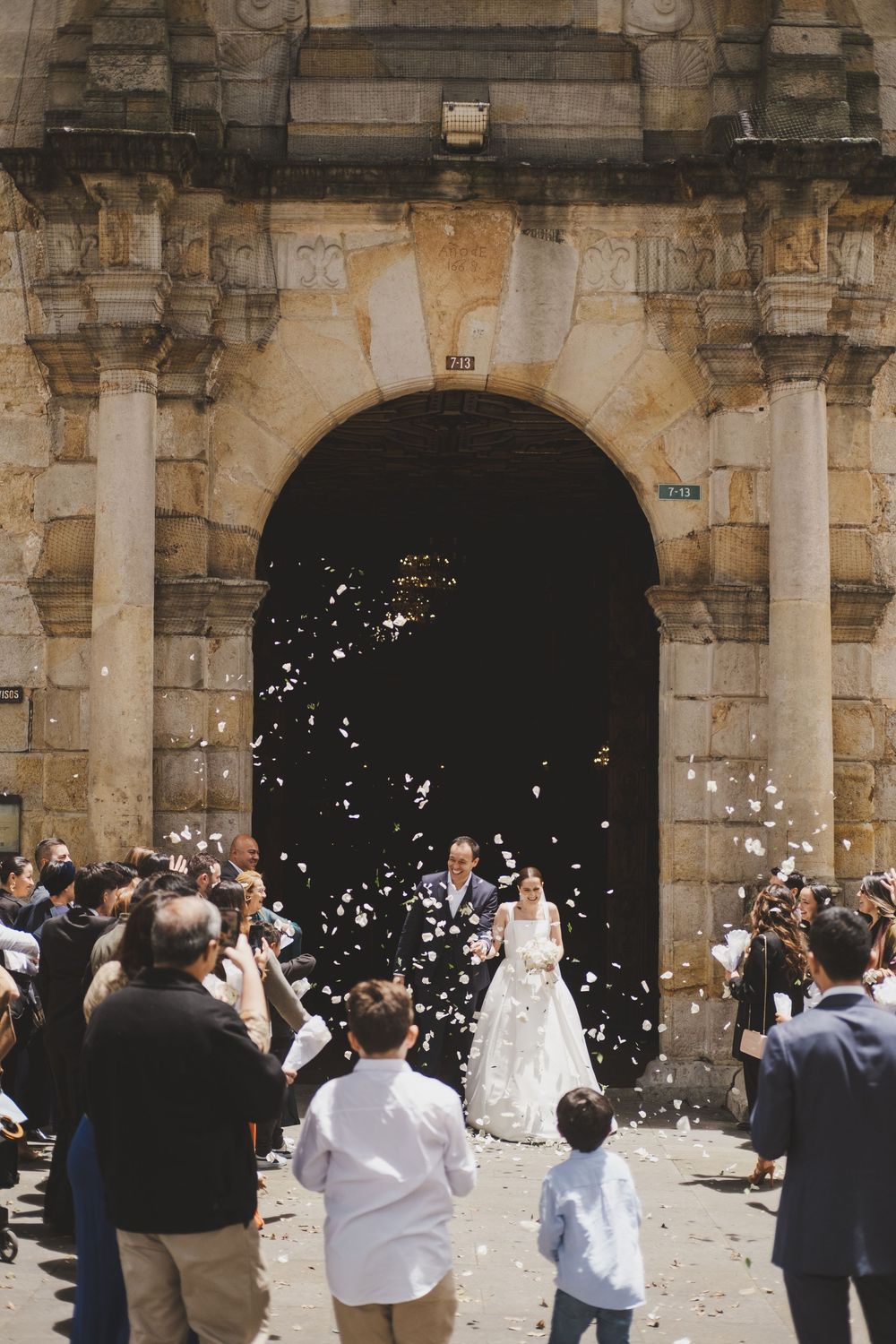 Wedding guests shower newlyweds with confetti under a grand stone archway entrance.