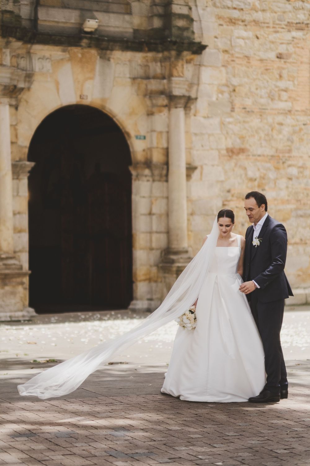 A romantic wedding moment captured in front of a historic stone archway with a flowing white dress and long veil.