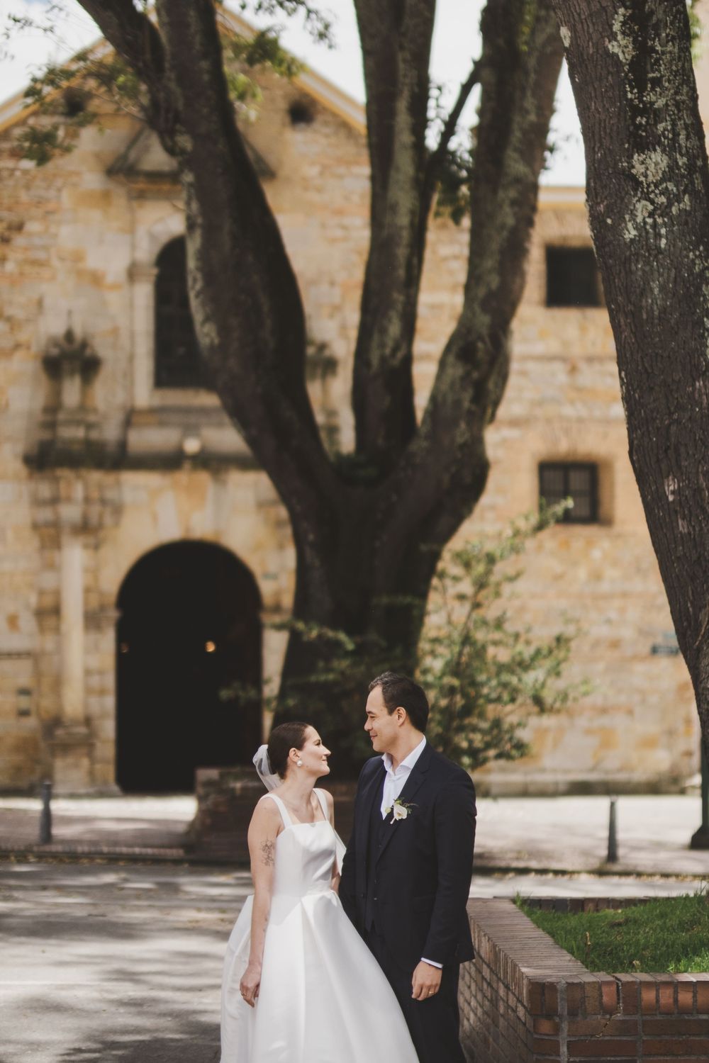 A couple in formal wedding attire share a moment outside a historic stone church building with arched doorways.