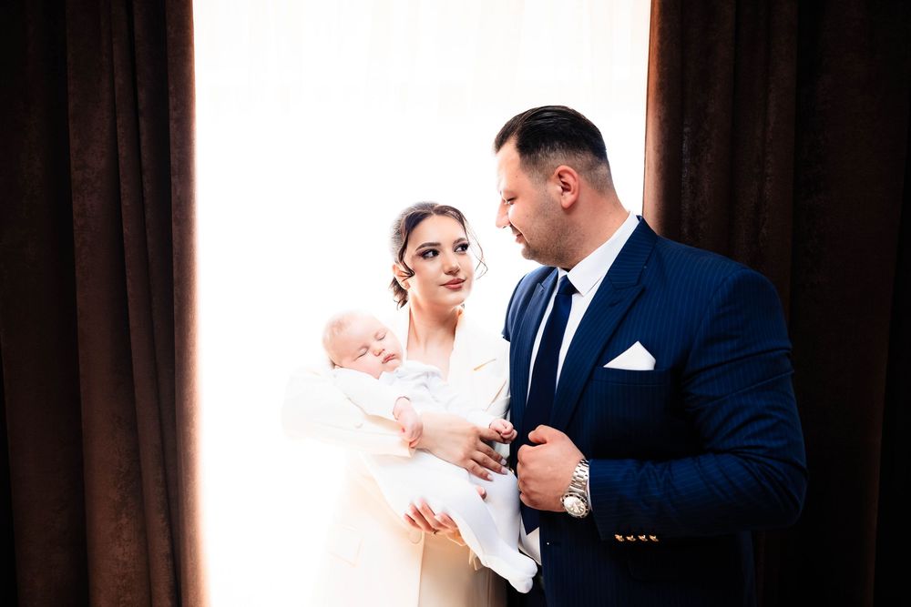 Elegant couple in formal attire holding their newborn baby near a window.