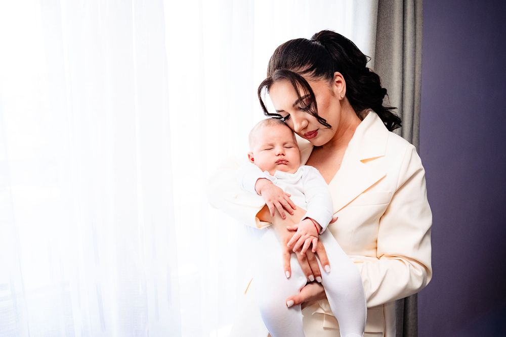 Woman in white blazer holding and gazing lovingly at a newborn baby by a window.