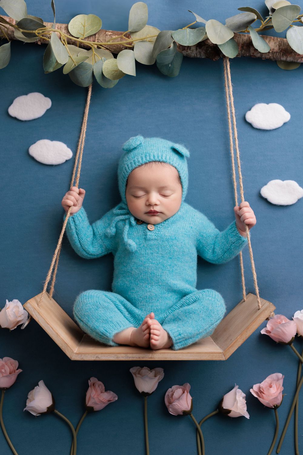A sleeping newborn in turquoise outfit rests on a wooden swing with floral decorations and paper clouds in the background.