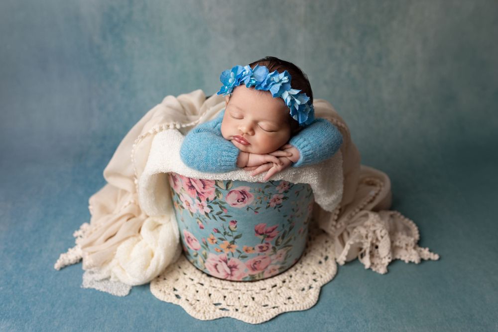 Newborn baby sleeping peacefully in vintage blue floral tea cup with lace backdrop and matching blue headpiece.