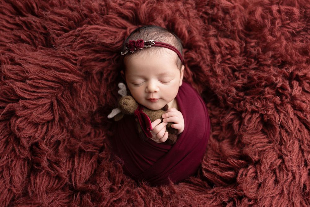 Sleeping newborn baby wrapped in burgundy swaddle with headband lying on fluffy red textured blanket.
