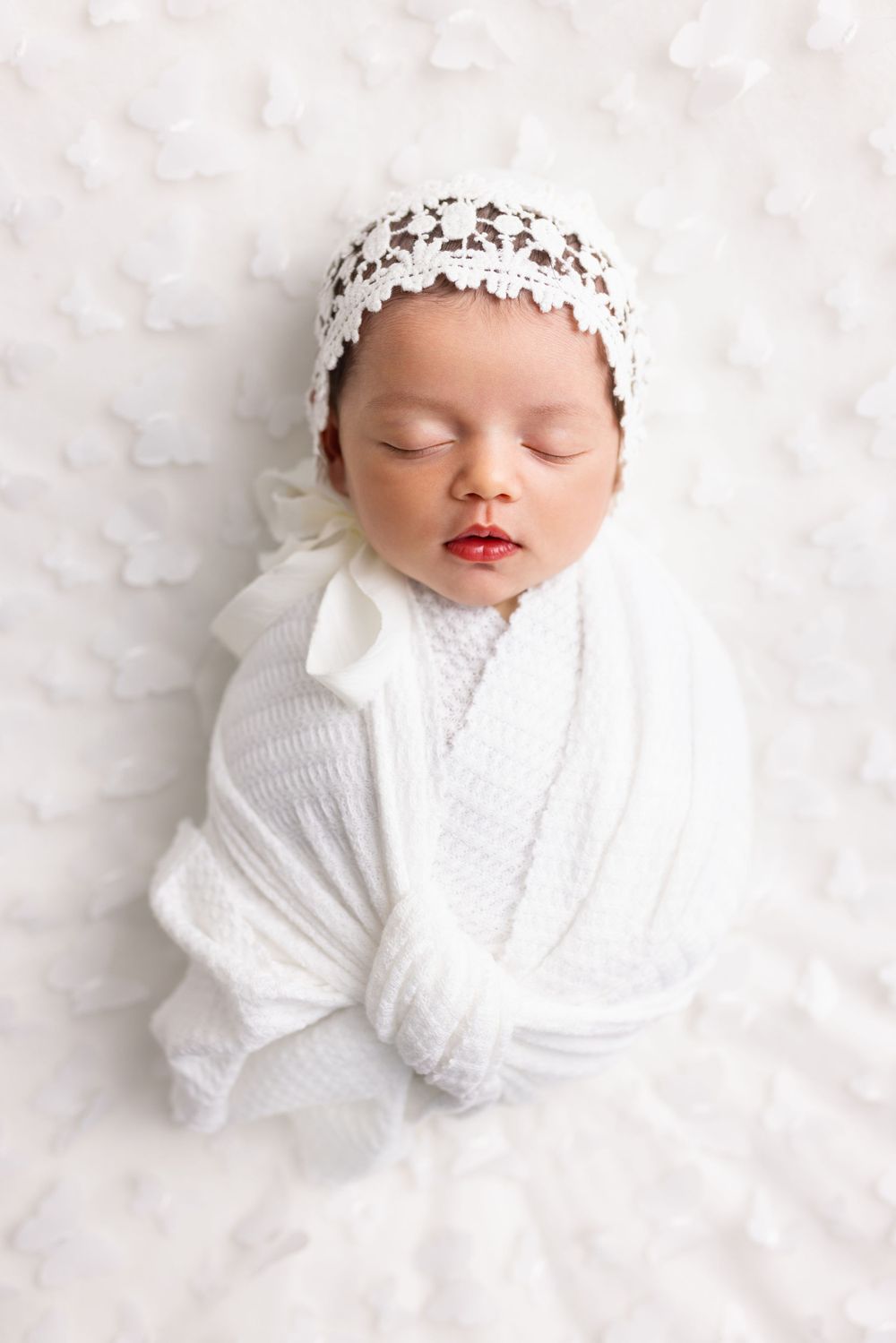 A sleeping newborn wrapped in white fabric wearing a delicate lace bonnet on a soft white background.