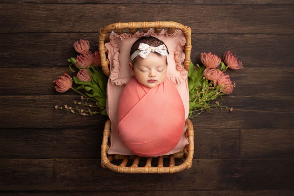 Newborn baby wrapped in coral pink swaddle sleeps in wicker basket surrounded by pink flowers on wooden backdrop.