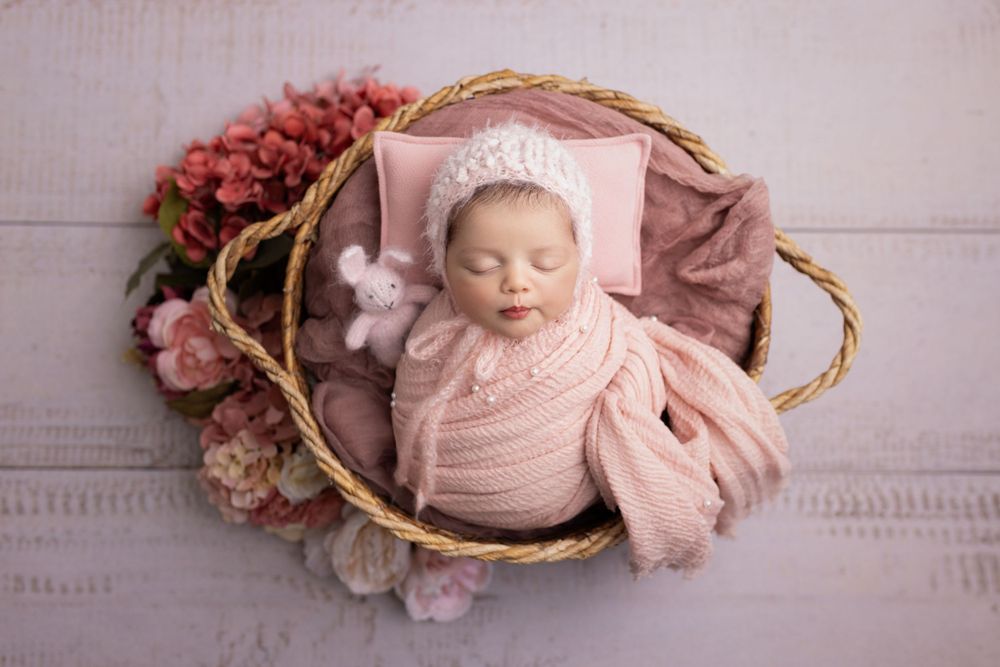 A sleeping newborn wrapped in pink swaddle cloth lies in a rustic basket adorned with pink and mauve flowers.