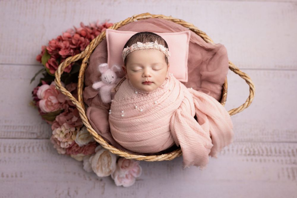 Sweet newborn sleeping peacefully in wicker basket surrounded by soft pink flowers and wrapped in rosy swaddle.