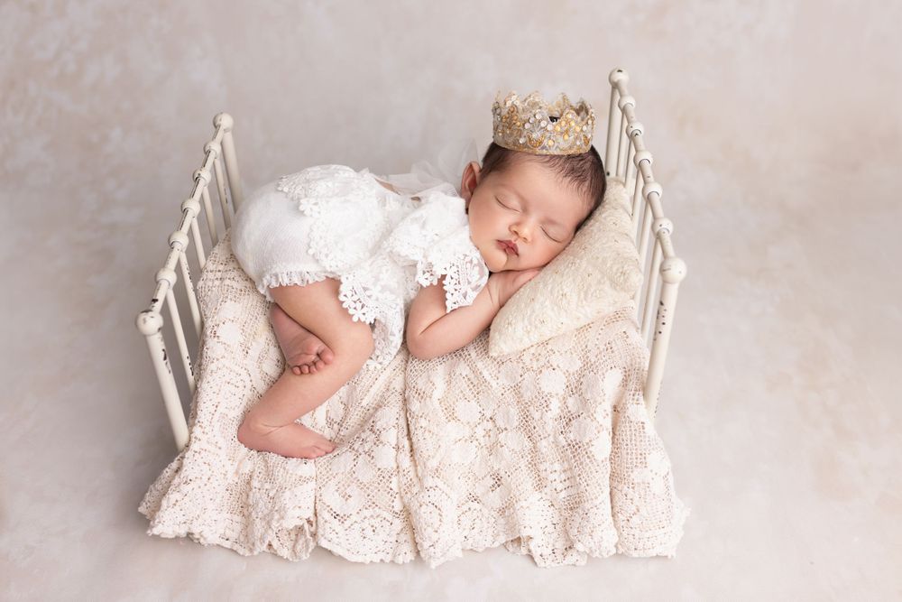 Newborn in white lace dress sleeps peacefully on delicate vintage white bed with crown accessory.
