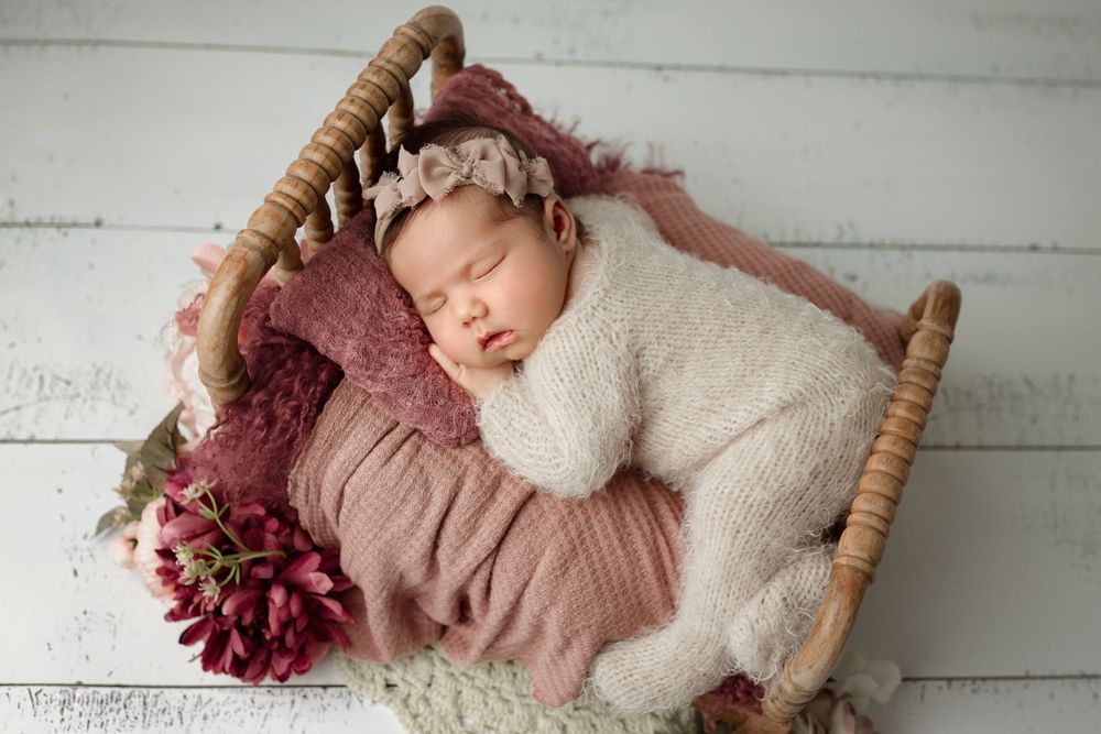 A sleeping newborn wrapped in a cozy white swaddle rests in a rustic basket with burgundy accents and floral decor in cedar park tx studio
