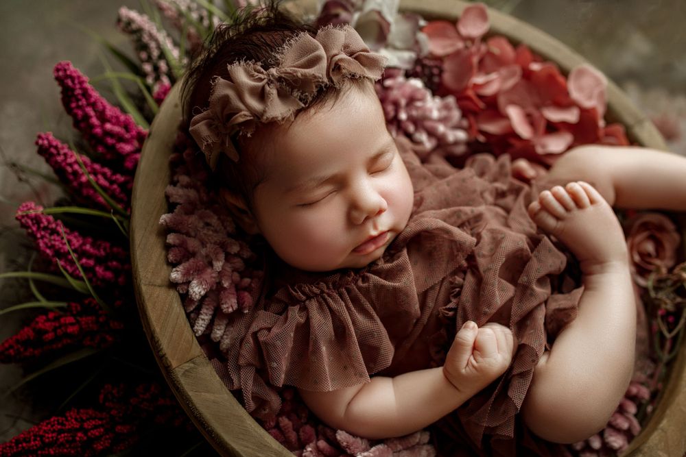 A sleeping baby in brown ruffled dress lies in a rustic wooden bowl with burgundy floral accents during newborn photos in cedar park tx