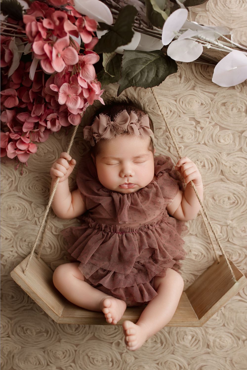 Newborn baby in a brown ruffled dress sleeping peacefully on a wooden swing with pink hydrangeas and white orchids nearby.