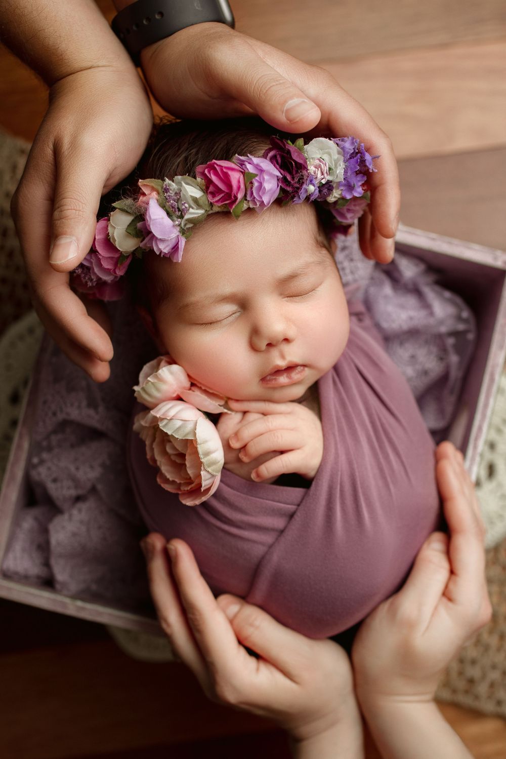 A sleeping baby wrapped in purple fabric wearing a floral crown of pink and white roses is held gently in caring hands during newborn photos