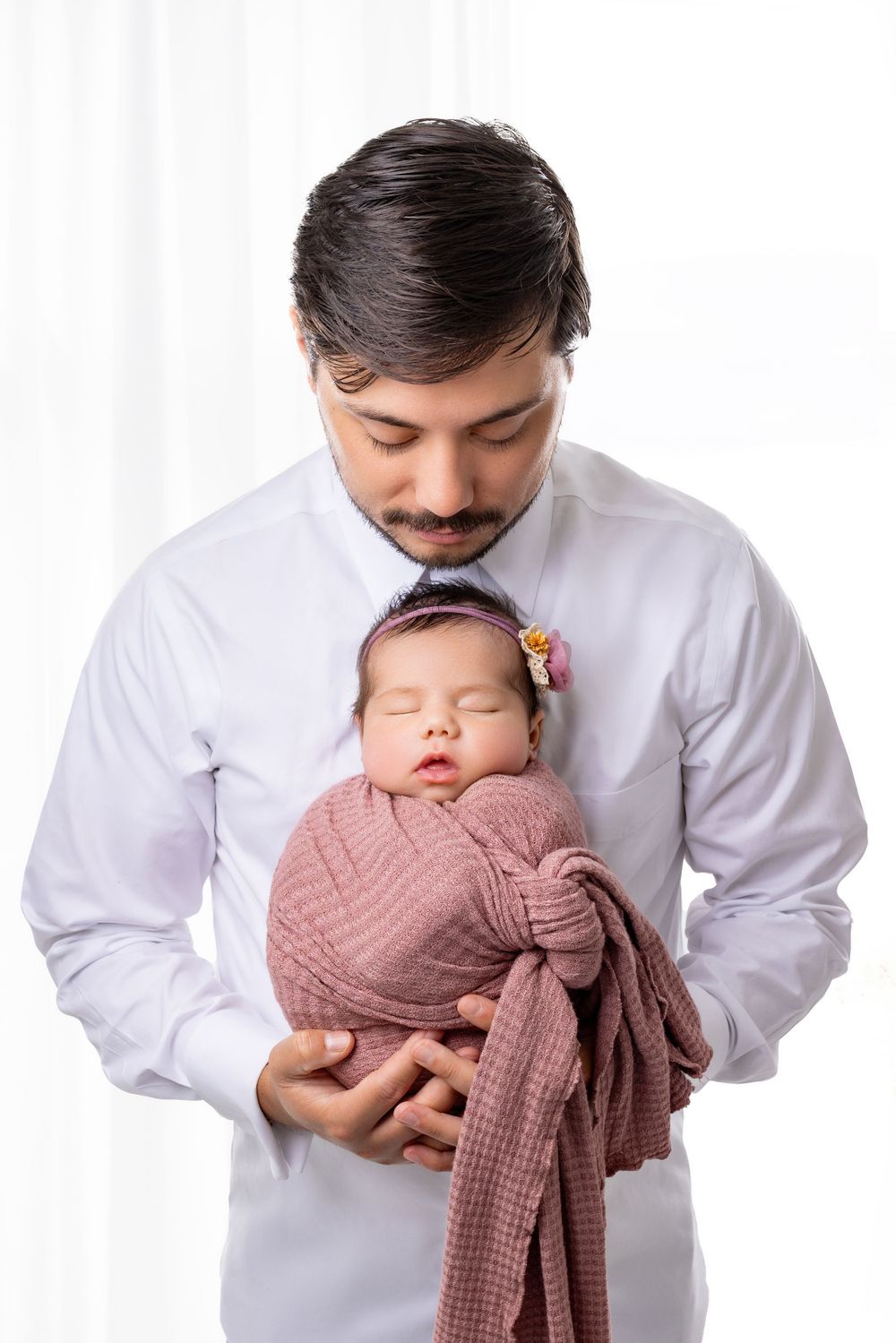 A tender moment captured as a sleeping newborn swaddled in pink is cradled gently against a father in a white button-up shirt, in Austin TX