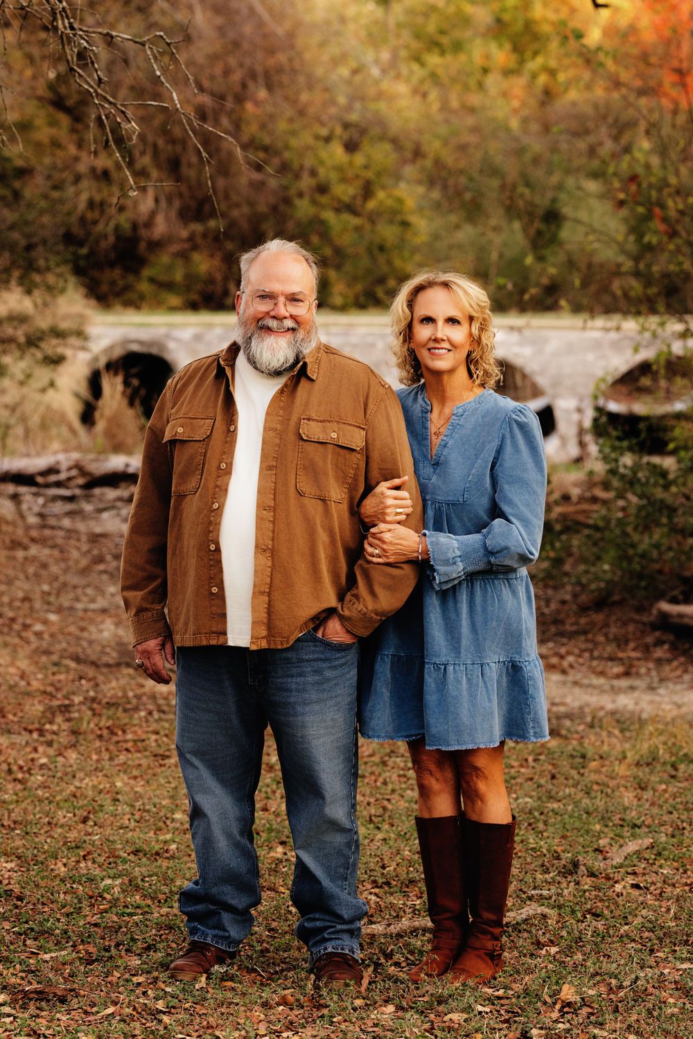 A couple stands together outdoors in fall, wearing casual attire with brown and blue clothing against an autumn backdrop.