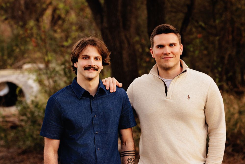 Two men standing shoulder to shoulder outdoors, one wearing a dark blue polo and mustache, other in beige pullover.