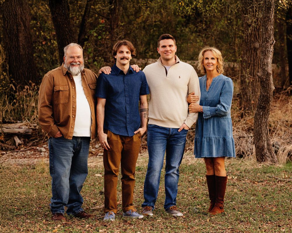 Family of four poses together outdoors during autumn with trees and fallen leaves in the background.